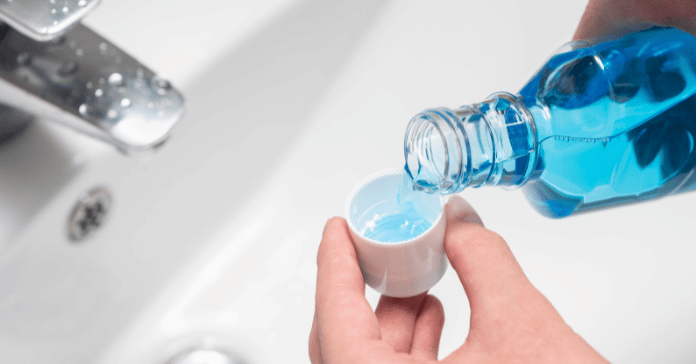 Hand pouring blue mouthwash into a cap over a bathroom sink for oral rinsing.